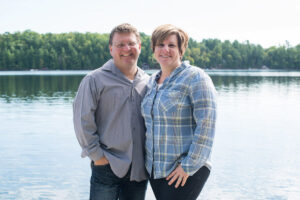 A photograph of two people, standing in front of a scenic mountain and lake background.